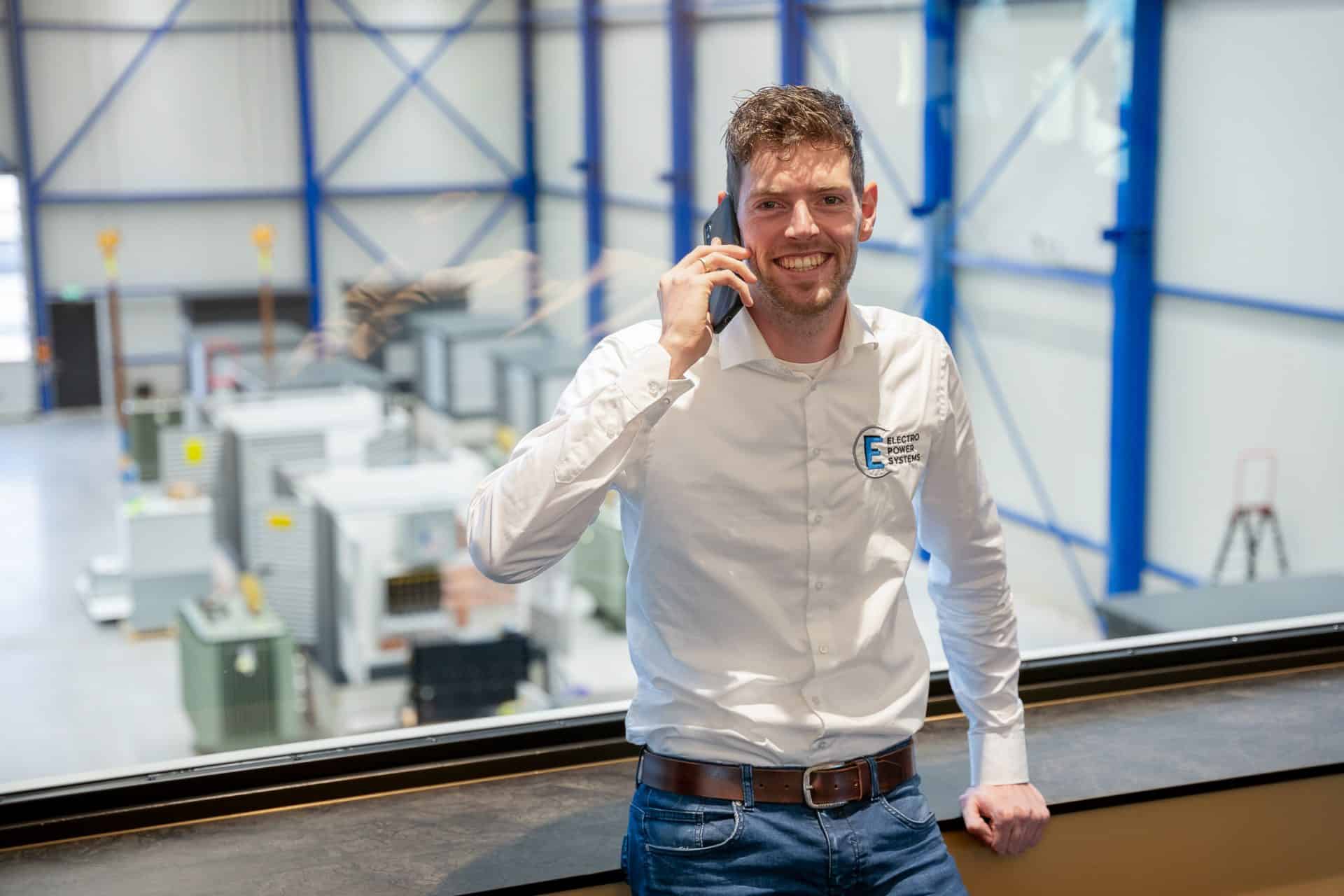 Een man in een wit overhemd en jeans staat bij een raam in een industriële omgeving, pratend op een mobiele telefoon en lachend. De achtergrond toont machines en apparatuur.