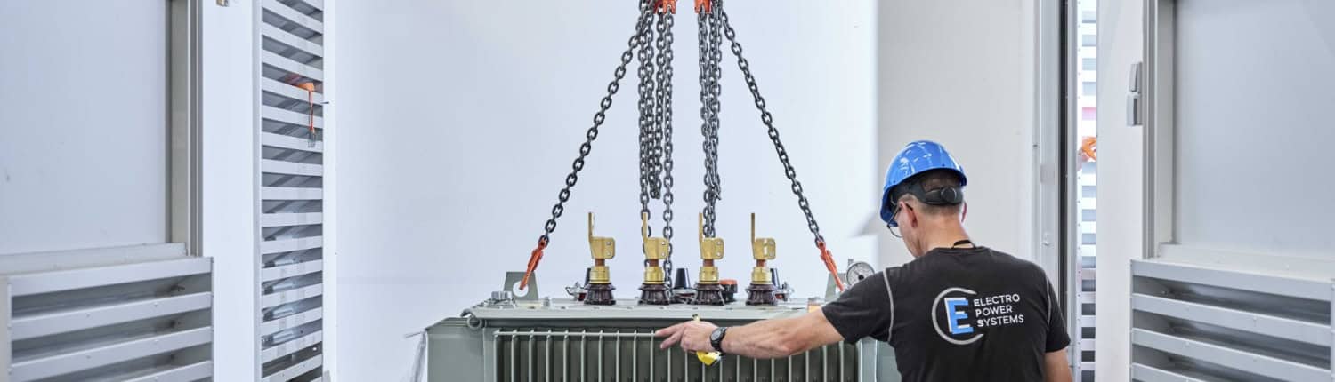 Een arbeider met een blauwe helm bedient een kraan om een transformator in een zeecontainer te laten zakken.