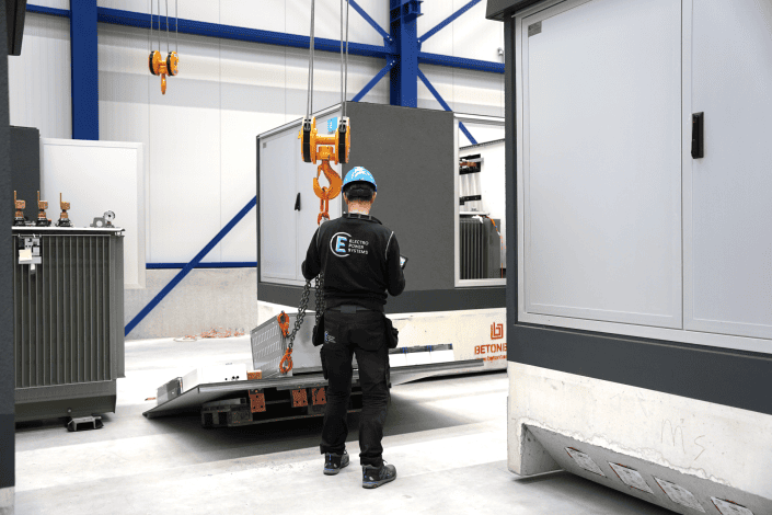Een arbeider in helm en uniform bedient een hijswerktuig om een grote, rechthoekige industriële unit in het magazijn te verplaatsen. Het doet denken aan de drukke taferelen die te zien zijn in ons aankomende nieuwsbericht van juli en augustus 2024.