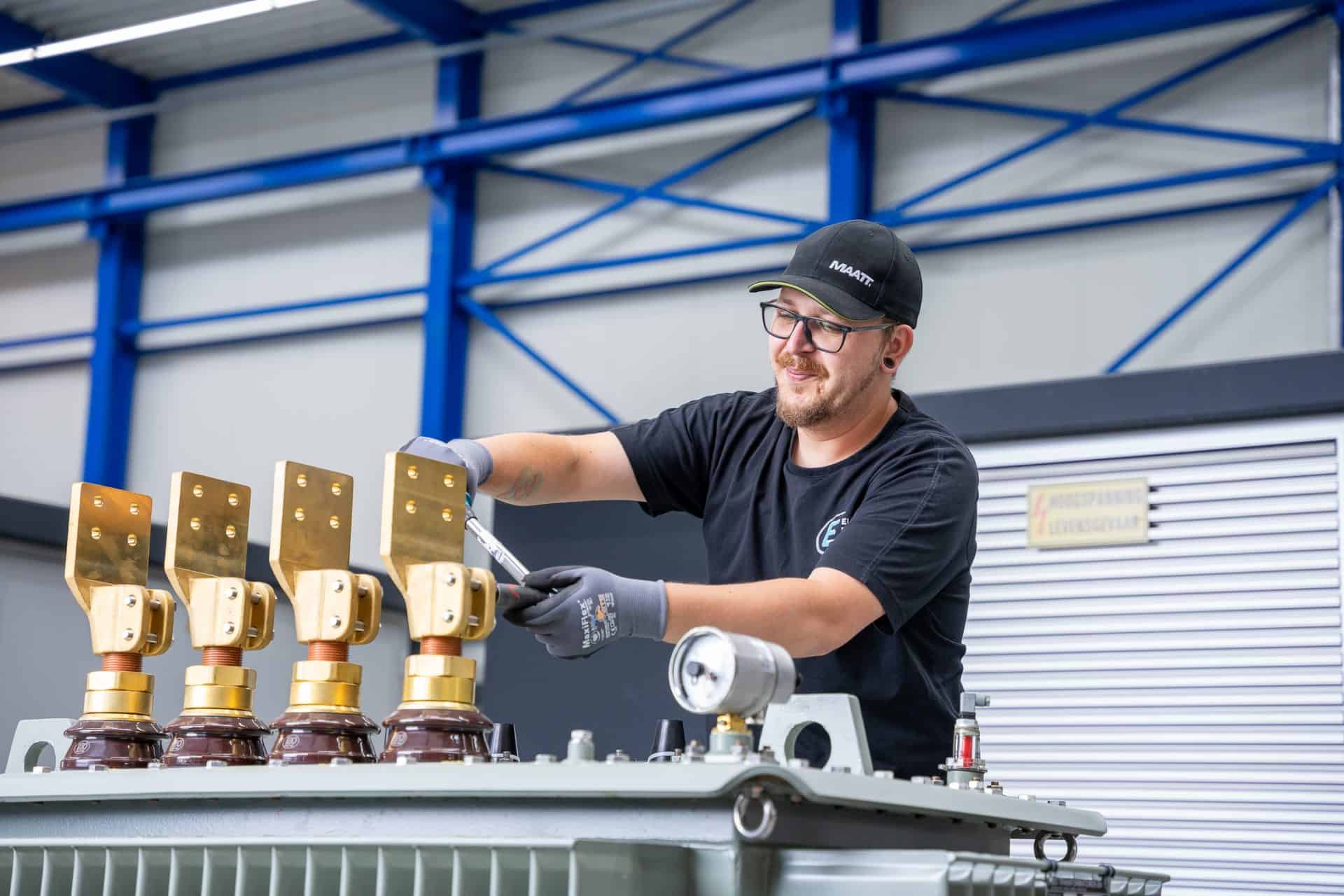 Een persoon draagt een zwarte pet en handschoenen en werkt in een magazijnachtige omgeving aan industriële apparatuur met metalen onderdelen.