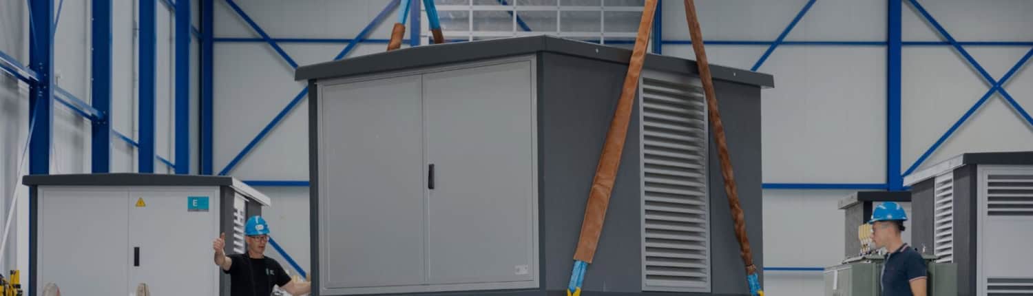 Twee arbeiders begeleiden een groot transformatorhuisje dat door een kraan in een magazijn wordt gehesen. Ze dragen helmen en beschermende kleding.