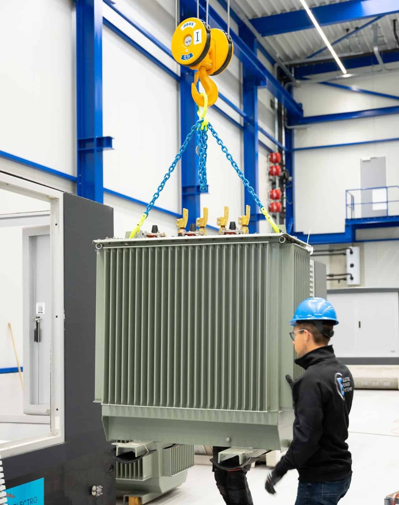 Een arbeider met een blauwe helm houdt toezicht op het hijsen van een grote grijze transformator met behulp van een gele kraan in een industriële faciliteit.