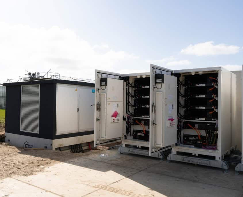 Rijen open batterijopslagunits en een gesloten schakelkast staan op een betonnen plaat, onder een heldere hemel, naast een grasveld. In productie: zes indrukwekkende stations voor Privatis Infrabeheer Zwolle verbeteren het landschap met technische precisie.