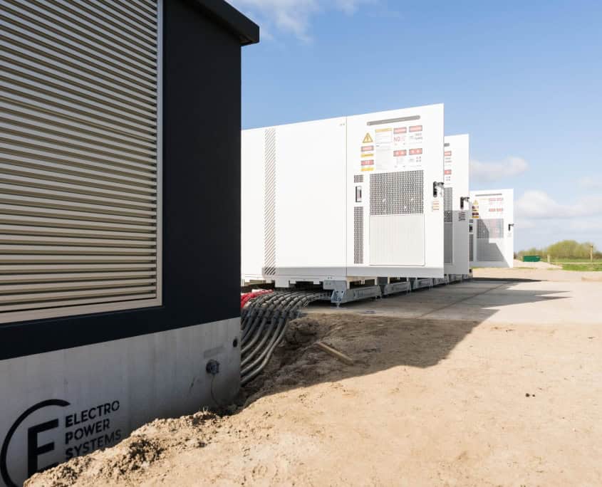 Buitenkant van het gebouw met meerdere grote witte industriële units op betonnen funderingen, omgeven door zand en gras, onder een strakblauwe lucht. Het toont indrukwekkende stations voor Privatis Infrabeheer Zwolle.