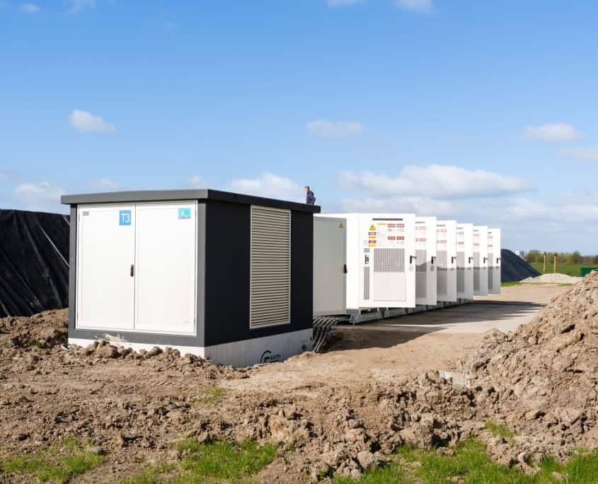 Een rij grote witte elektrische units, onderdeel van de ondergrondse stations voor Privatis Infrabeheer Zwolle, omzoomt een onverhard pad onder een blauwe lucht met hier en daar wat wolken.