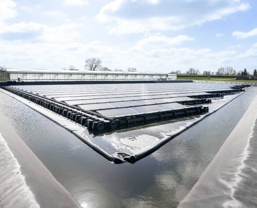 Drijvende zonnepanelen op een groot waterreservoir onder een deels bewolkte hemel, omgeven door groene velden en een verre kas, symboliseren de innovatie in de zes indrukwekkende stations van Privatis Infrabeheer Zwolle.
