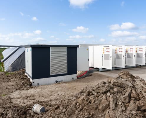 In productie: zes indrukwekkende stations voor Privatis Infrabeheer Zwolle, worden verschillende grote batterijopslageenheden en infrastructuur buiten op een betonnen plaat geïnstalleerd, omgeven door aarde en gras onder een gedeeltelijk bewolkte hemel.