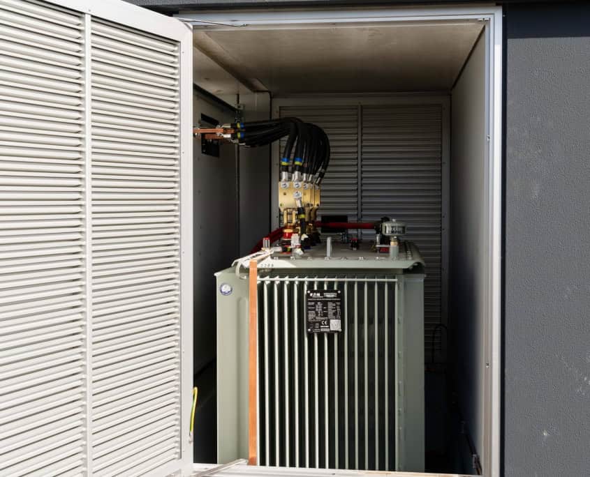 Een transformatorunit in een gedeeltelijk open metalen behuizing, met zichtbare kabels en connectoren, maakt deel uit van de indrukwekkende stations die momenteel in productie zijn voor Privatis Infrabeheer Zwolle.