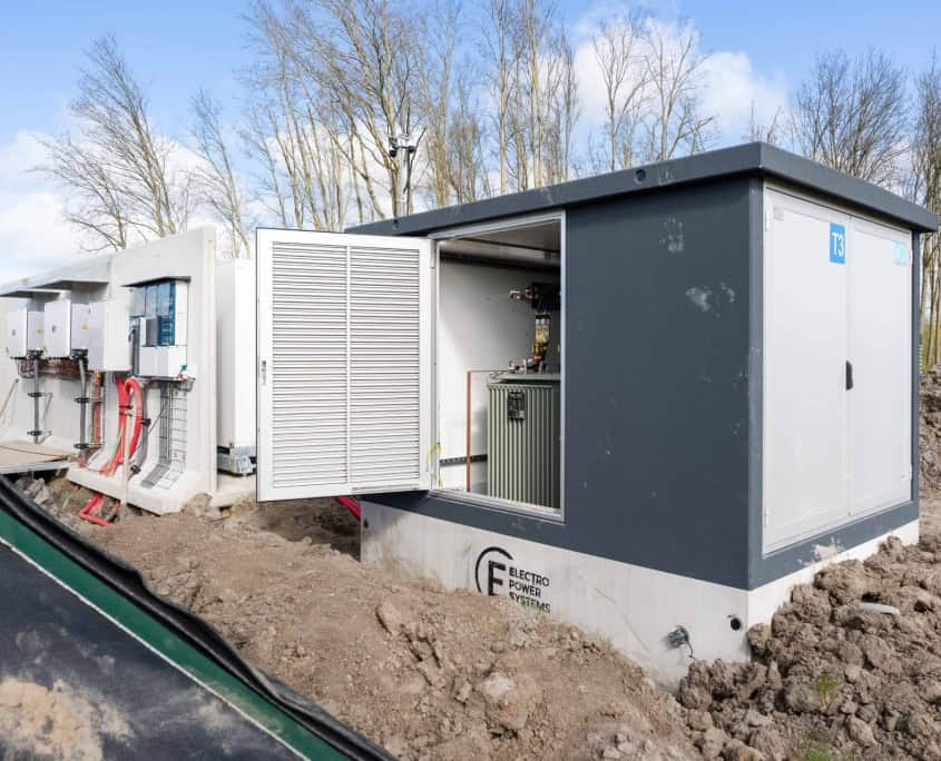 Een rij elektrische onderstations met open deuren die hun ingewikkelde interne componenten onthullen, staat op een modderige bouwplaats tussen bladerloze bomen. In productie: zes indrukwekkende stations voor Privatis Infrabeheer Zwolle, die de vooruitgang van de moderne infrastructuur weerspiegelen.