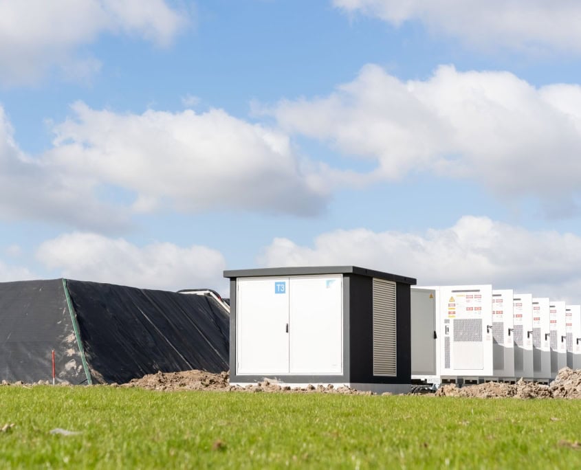 Een rij witte elektrische units, zes indrukwekkende stations in productie voor Privatis Infrabeheer Zwolle, staat opgesteld naast een met een zeil bedekte heuvel onder een blauwe lucht met hier en daar wat wolken.