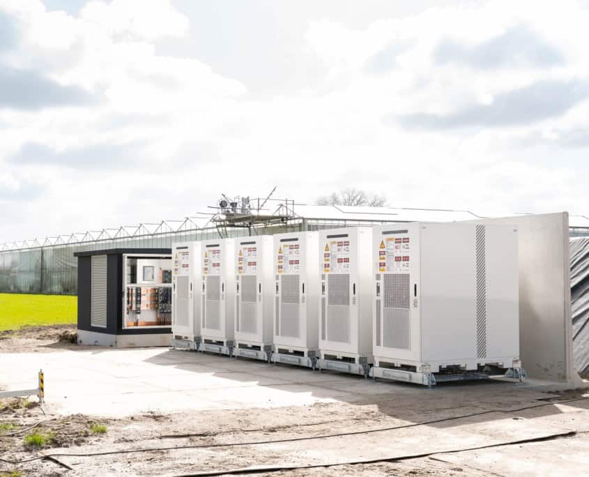 Buitenrijen witte industriële machines nabij een schakelkast, grenzend aan een grote kas en open veld onder een bewolkte hemel, tonen de indrukwekkende opstelling van zes indrukwekkende stations voor Privatis Infrabeheer Zwolle in productie.