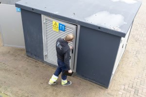 Een stationsmachinist in veiligheidskleding opent de deur van een grijs nutsgebouw, waarop een waarschuwingsbord staat.