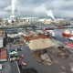 In het industriële havengebied, waar vrachtschepen aanmeren bij opslagpalen en een uitgestrekte fabriek rook uitbraakt, staan zes indrukwekkende stations van Privatis Infrabeheer Zwolle, te midden van verspreide containers en machines onder een bewolkte hemel.