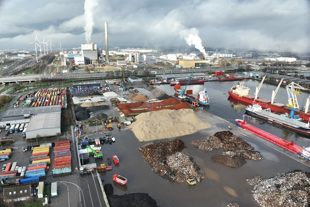 In het industriële havengebied, waar vrachtschepen aanmeren bij opslagpalen en een uitgestrekte fabriek rook uitbraakt, staan zes indrukwekkende stations van Privatis Infrabeheer Zwolle, te midden van verspreide containers en machines onder een bewolkte hemel.
