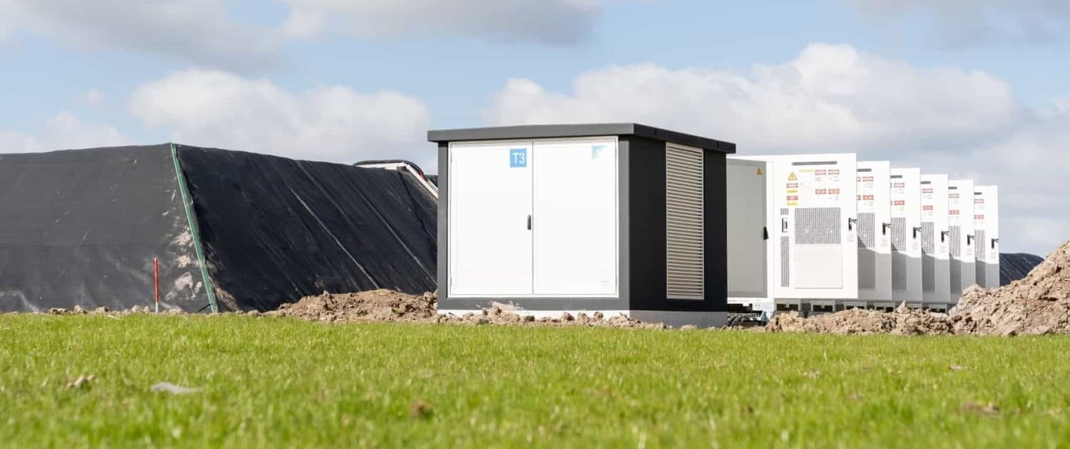 Een rij witte elektriciteitskasten staat op een grasveld, met op de achtergrond hopen aarde en een bedekte heuvel onder een gedeeltelijk bewolkte hemel.