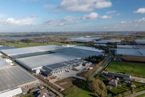 Luchtfoto van grote kassencomplexen, landbouwgrond en landelijke gebouwen onder een gedeeltelijk bewolkte hemel, met een transformatorstation dat de agrarische sector ondersteunt en windturbines zichtbaar in de verte.