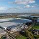 Luchtfoto van grote kassencomplexen, landbouwgrond en landelijke gebouwen onder een gedeeltelijk bewolkte hemel, met een transformatorstation dat de agrarische sector ondersteunt en windturbines zichtbaar in de verte.