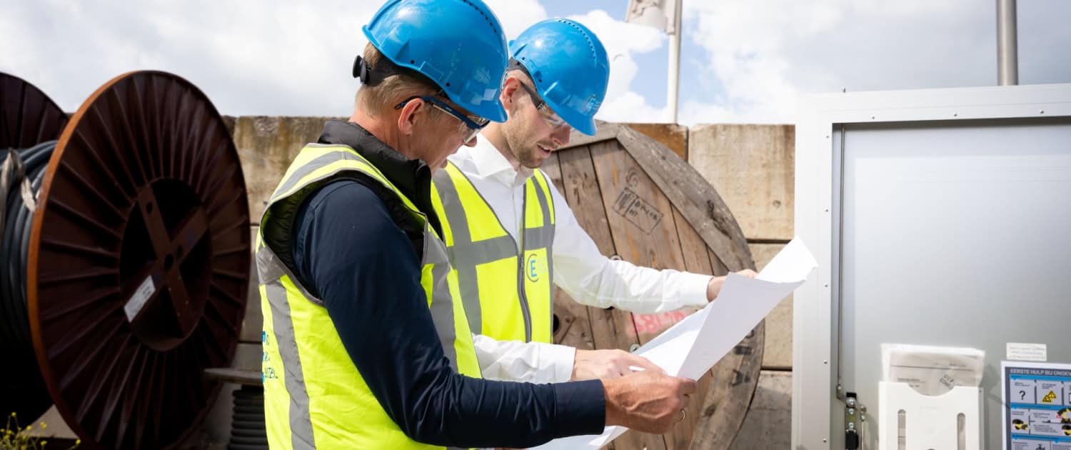 Twee bouwvakkers met blauwe helmen en gele veiligheidsvesten bekijken blauwdrukken op een buitenwerkplek in de buurt van kabelhaspels en apparatuur van Electro Power Systems.