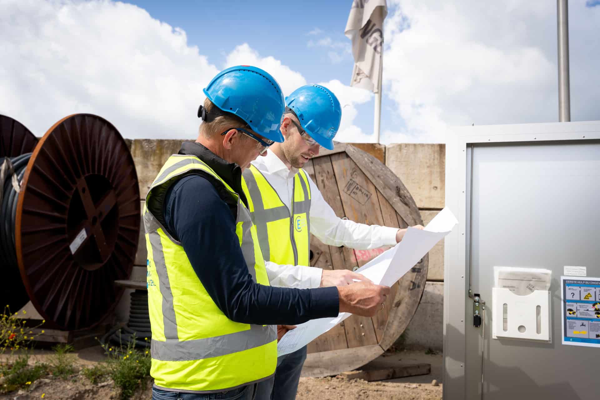 Twee bouwvakkers met blauwe helmen en gele veiligheidsvesten bekijken blauwdrukken op een buitenwerkplek in de buurt van kabelhaspels en apparatuur van Electro Power Systems.