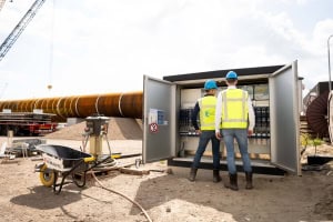 Twee arbeiders in veiligheidsuitrusting staan bij een open bedieningspaneel van Electro Power Systems op een bouwterrein, met grote pijpen en apparatuur op de achtergrond.