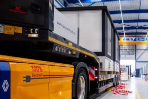Een 100% elektrische vrachtwagen met aanhanger staat geparkeerd in een industrieel magazijn, met een groot stuk apparatuur voor duurzame transformatorstations.