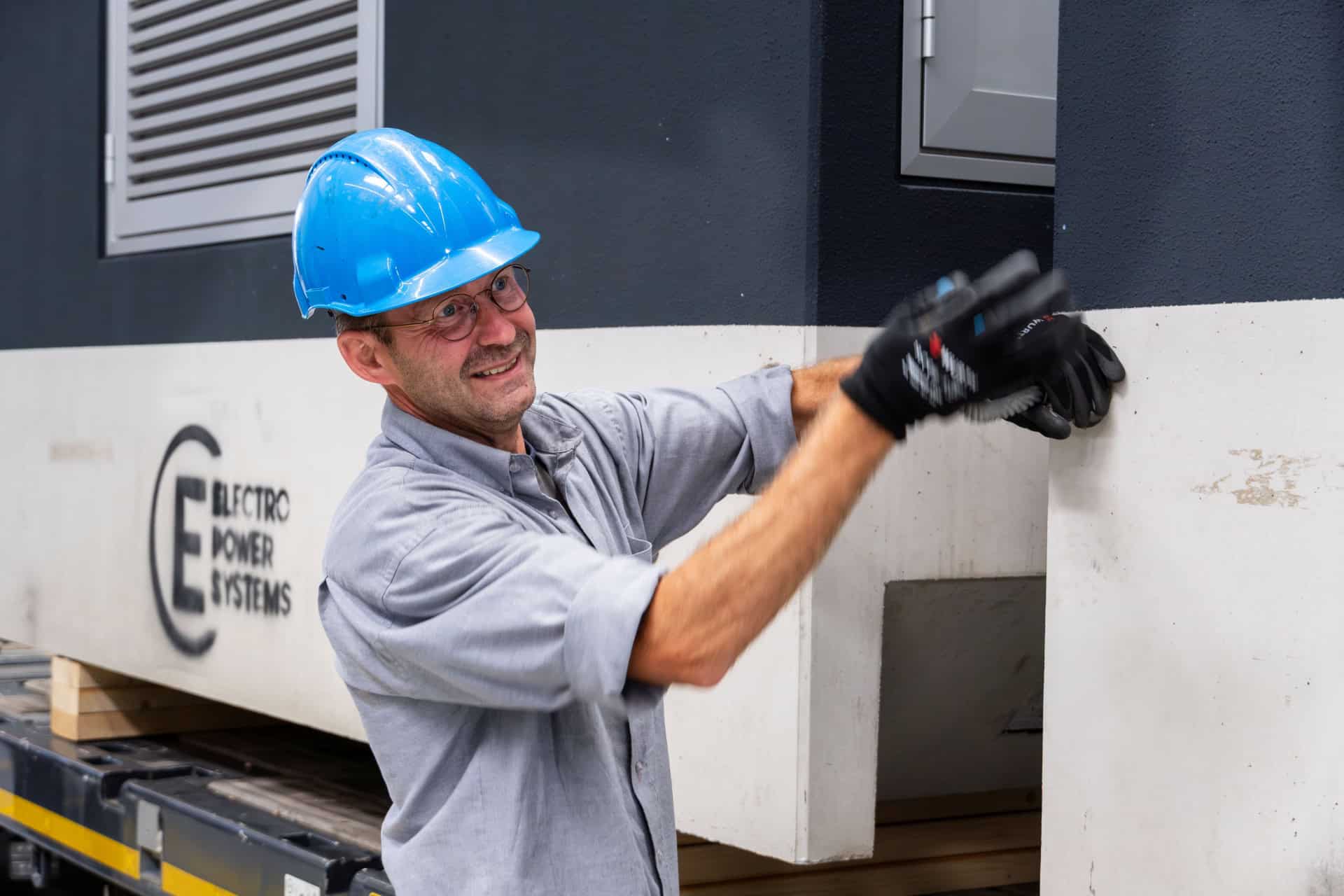 Een arbeider met een blauwe helm en handschoenen installeert of inspecteert apparatuur bij een structuur met het label "Electro Power Systems," die bijdraagt aan duurzame transformatorstations voor efficiëntere en duurzamere energieoplossingen.