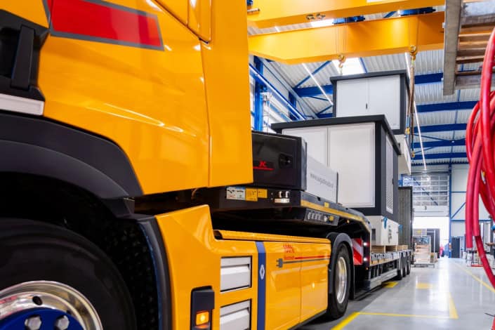 Een gele vrachtwagen vervoert industriële apparatuur, waaronder duurzame transformatorstations, in een ruim, goed verlicht magazijn met blauwe en witte interieuraccenten.