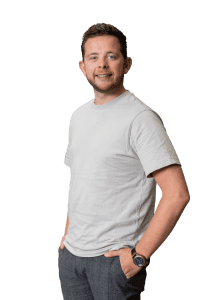 Een man met kort haar en een baard, in een lichtgrijs T-shirt en grijze broek, staat met zijn handen in zijn zakken tegen een effen witte achtergrond - perfect voor een Over ons introductie.