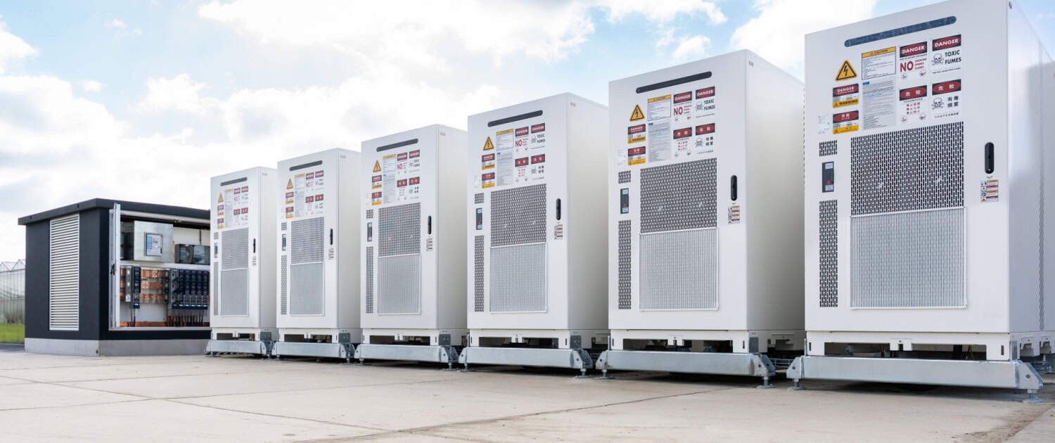 Een rij grote witte industriële elektriciteitskasten en een bedieningspaneelstructuur van een trafostation buiten geïnstalleerd op een betonnen ondergrond onder een gedeeltelijk bewolkte hemel.