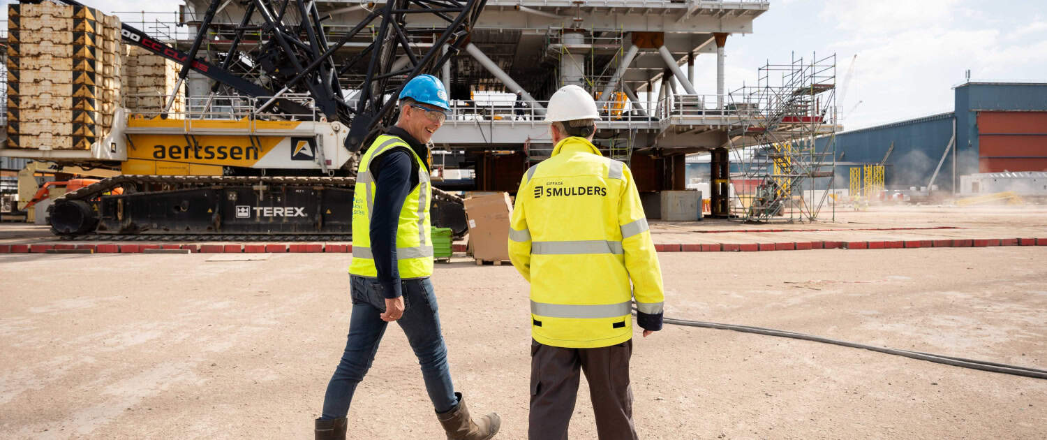 Twee bouwvakkers met veiligheidsuitrusting lopen op een bouwterrein met een grote kraan en een industrieel gebouw op de achtergrond.