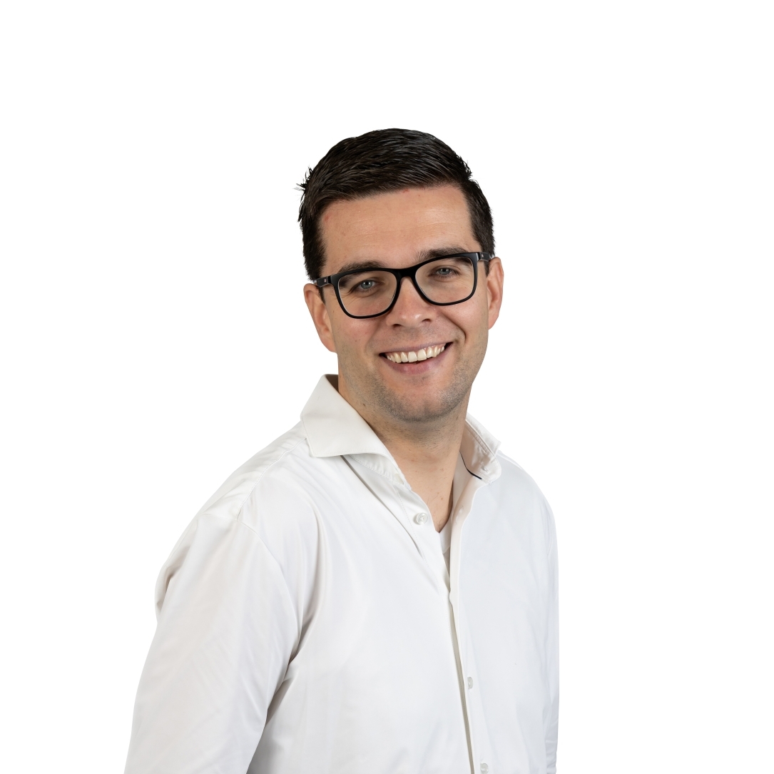 Man met bril, wit overhemd en donkere spijkerbroek staat met een hand in zijn zak en glimlacht zelfverzekerd tegen een effen witte achtergrond, de belichaming van de toegankelijke stijl van een Technisch Ingenieur.