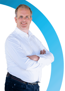 Man met kort licht haar draagt een wit overhemd met knoopsluiting en een donkere spijkerbroek staat glimlachend met gekruiste armen voor een witte en blauwe gebogen achtergrond.