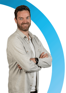 Een man met kort bruin haar en een baard staat met gekruiste armen, draagt een licht jack en smartwatch, voor een witte achtergrond met een blauwe gebogen afbeelding.