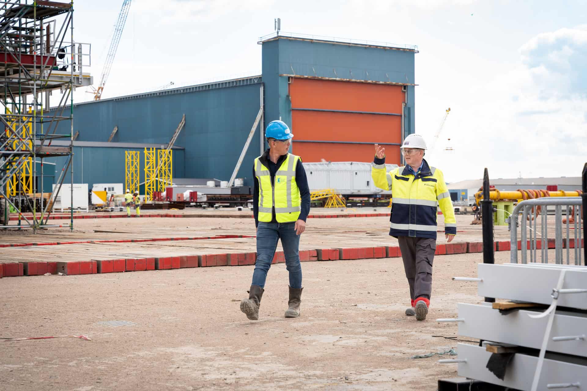 Twee mensen met veiligheidshelmen en reflecterende vesten lopen en praten op een bouwplaats, waar industriële gebouwen en apparatuur het nieuwe Snellaadstation Almelo voorzien van betrouwbare energie omringen.