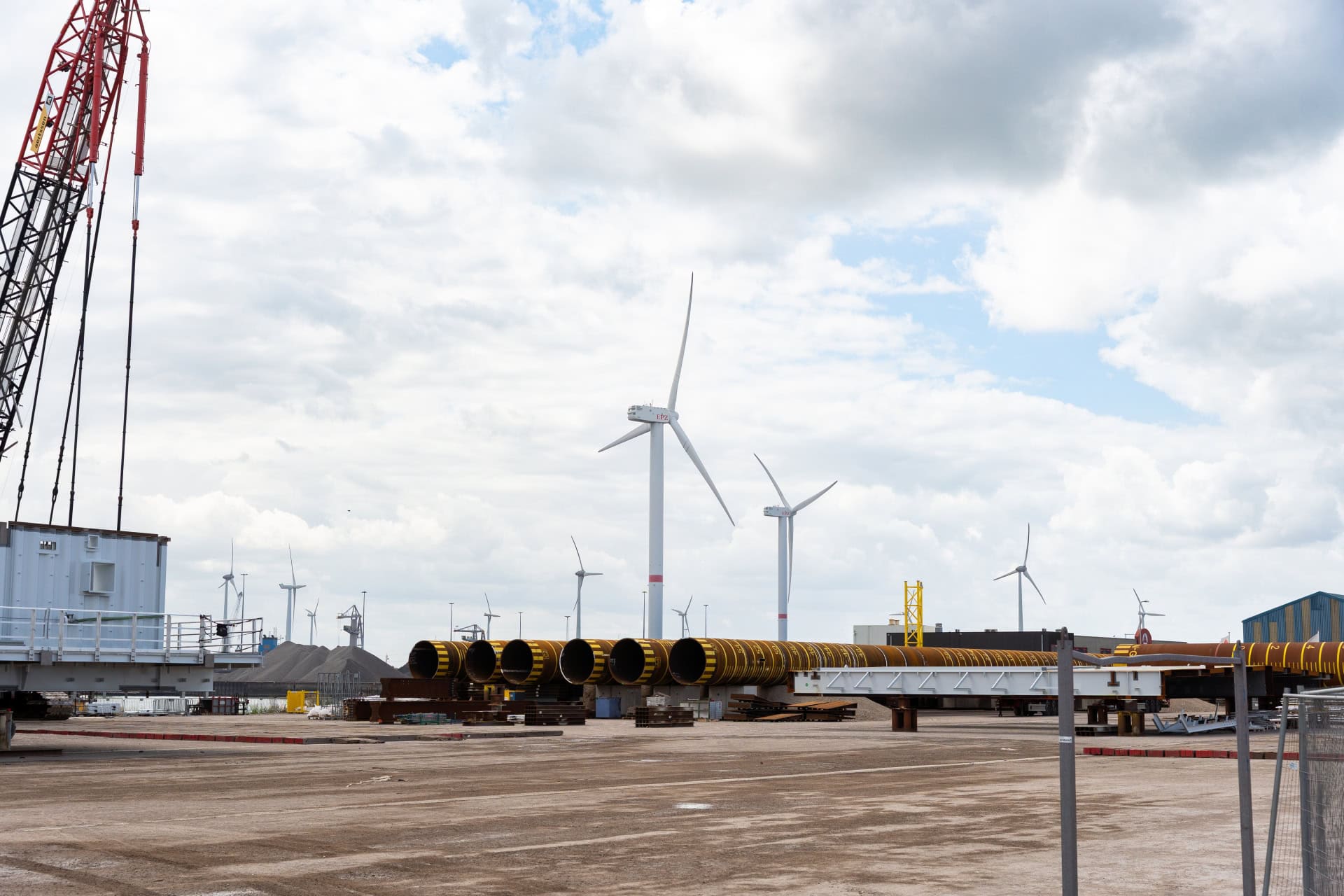 Windturbines staan op de achtergrond van een industrieterrein, waar grote gele pijpen en bouwmachines werken onder een bewolkte hemel-Snellaadstation Almelo voorzien van betrouwbare energie.