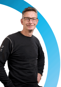 Een man met een bril en een zwarte trui staat met een hand op zijn heup voor een witte achtergrond met een blauwe gebogen afbeelding, die de professionele geest van onze pagina Over ons belichaamt.