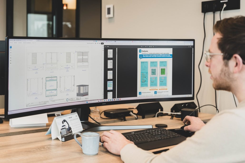 Een Projectleider 10kV werkt aan een bureau en gebruikt een computer met een ultrabreed beeldscherm om technische diagrammen en documenten weer te geven.