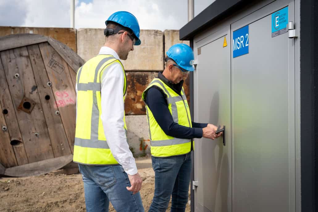Twee arbeiders in veiligheidsvesten en helmen staan bij een industriële kast met het label "NSR 2"; een Projectleider 10kV bedient het bedieningspaneel terwijl de andere toekijkt.