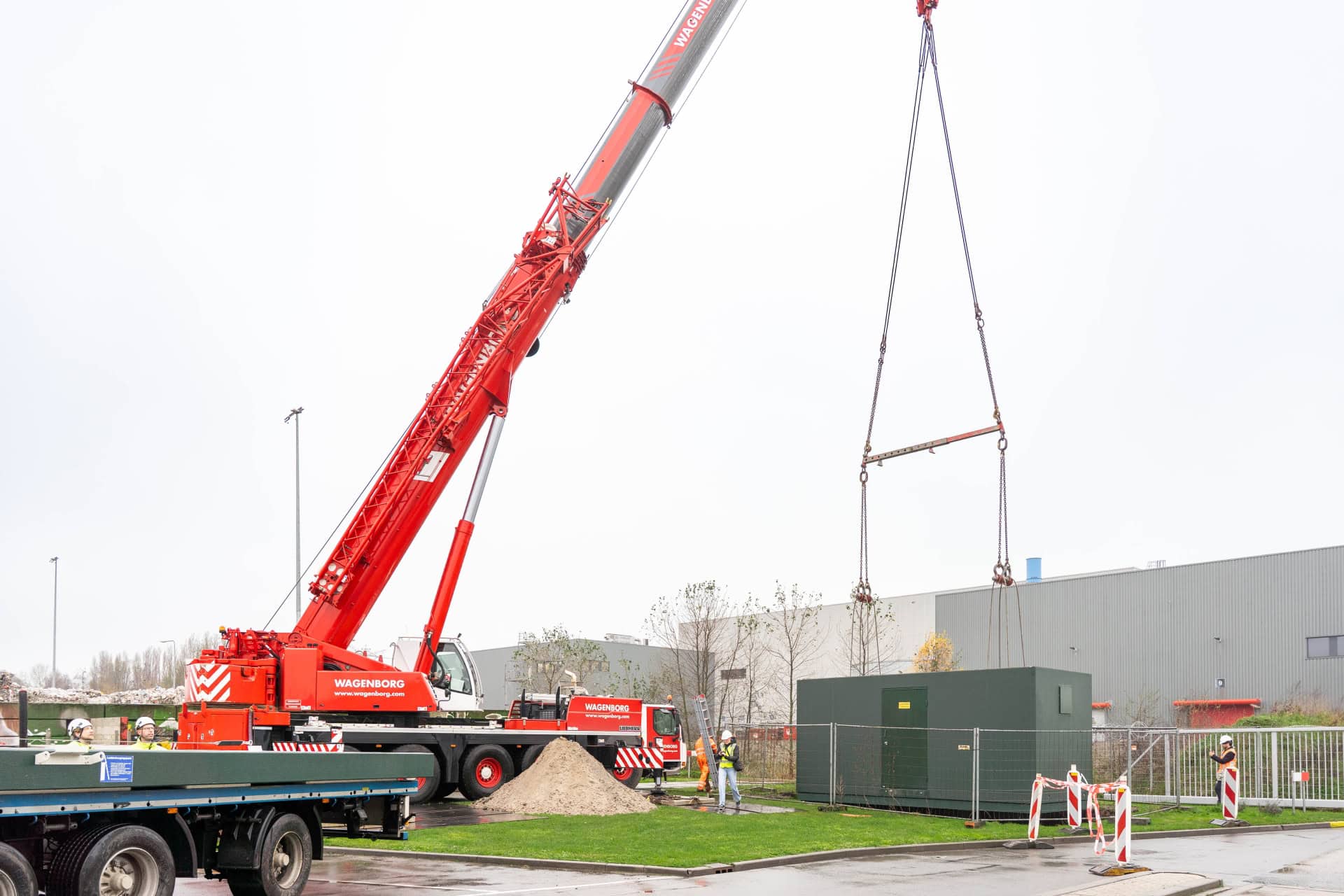 Een grote rode kraan tilt een groene industriële container op terwijl arbeiders in veiligheidsuitrusting toezicht houden op een bouwterrein bij een grijs gebouw, waar de energie achter 30.000 kilo paprika per dag wordt getoond.