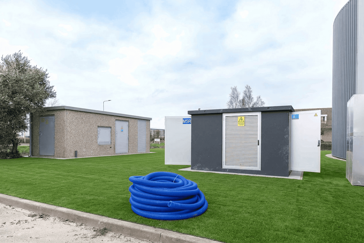 Een klein nutsgebouw en een elektrisch onderstation, onderdeel van negen transformatorstations voor offshore constructiewerkzaamheden, staan op een grasveld; een spoel van blauwe flexibele leiding ligt op de grond op de voorgrond.