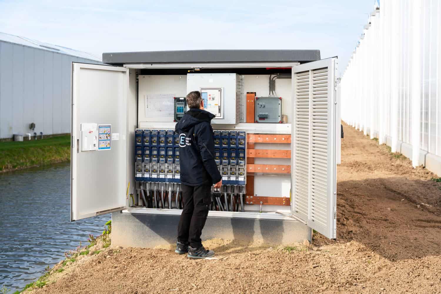 Een persoon staat voor een open elektrisch bedieningspaneel naast een kas op Ecopark De Wierde, waar de apparatuur voor het energie-uitbreidingsproject wordt geïnspecteerd.