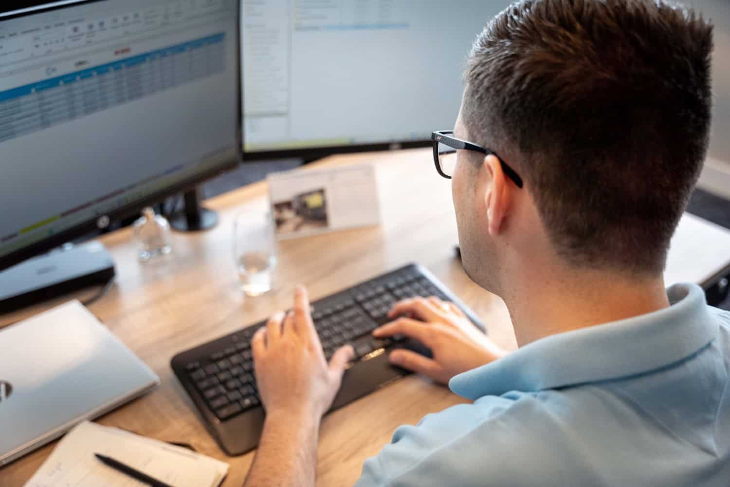 Een brildragende persoon zit achter een bureau met een toetsenbord en twee computermonitors en maakt aantekeningen in een notitieblok dat naast hem ligt – een voorbeeld van de werkomgeving bij Werken bij Electro Power Systems.